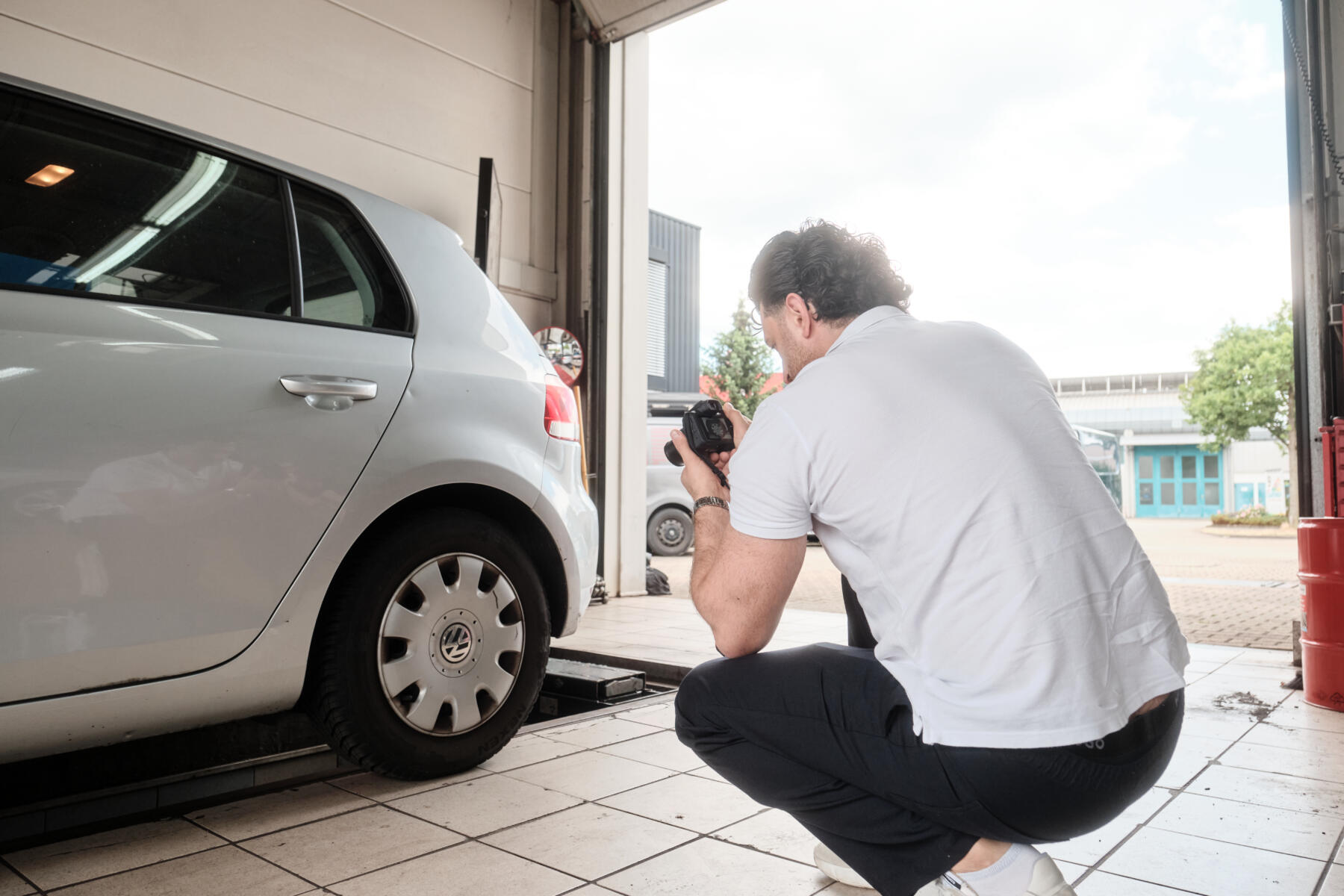 Ein Gutachter kniet neben einem grauen Auto und fotografiert einen Schaden in einer Werkstatt für die Dokumentation.